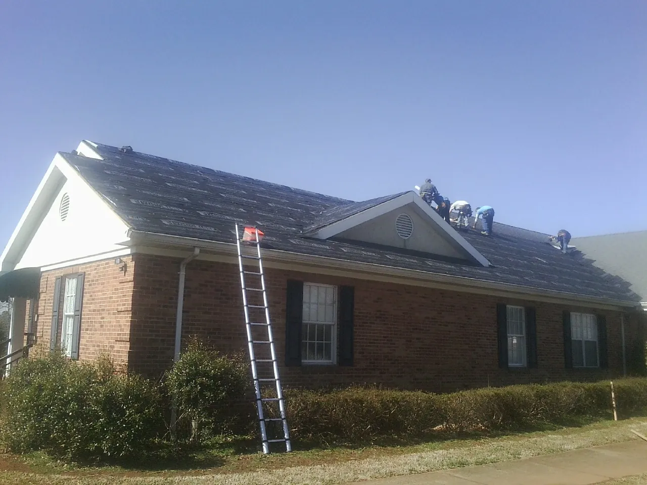Crew tearing shingles off a brick ranch roof with a ladder set — Emergency Roof Repair work by our crew in Metz