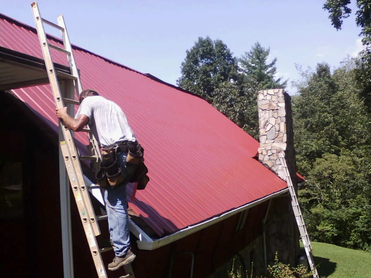 Roofer on a ladder beside a red metal roof and stone chimney — Soffit & Fascia Repair by Roofing Brothers in Metz